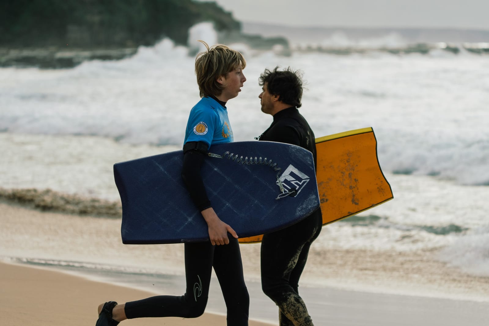 Two young riders carrying their bodyboards along the shore