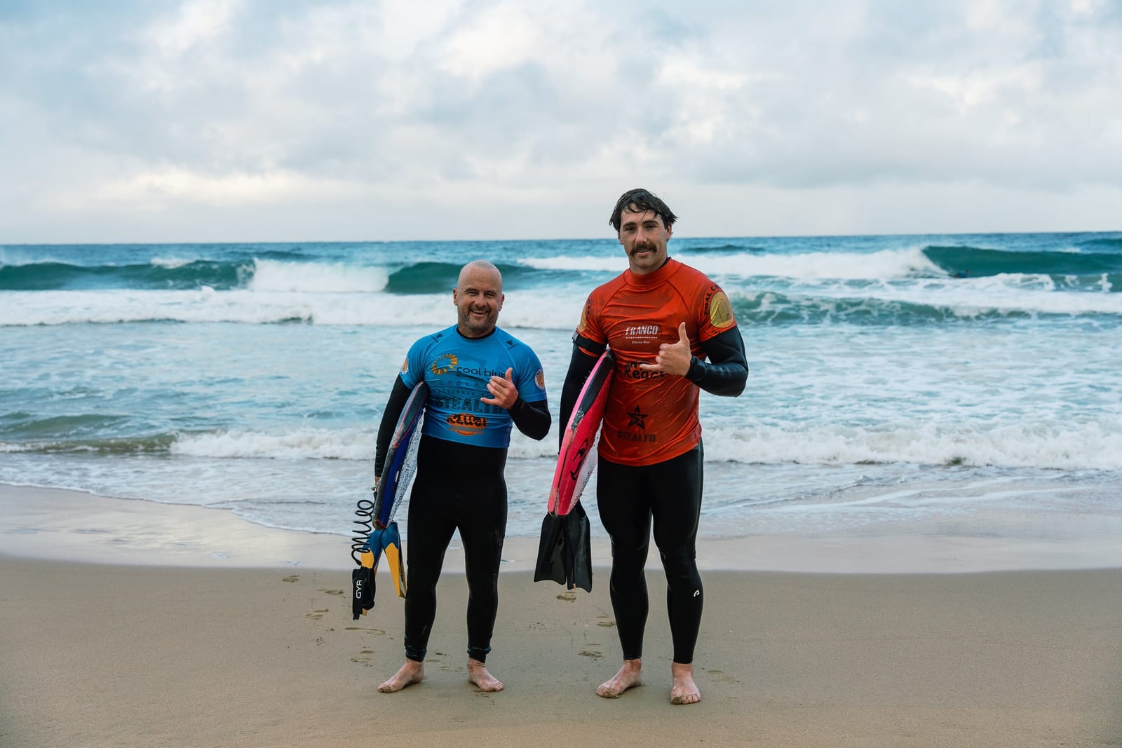 Two club members with their boards, smiling for the camera on the sand